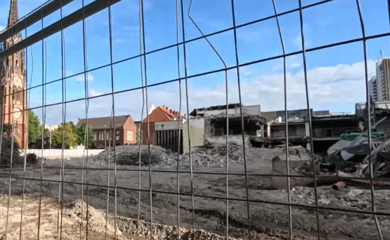 Abrissfläche des ehemaligen Karstadt-Gebäudes hinter einem Bauzaun, im Hintergrund die Große Kirche